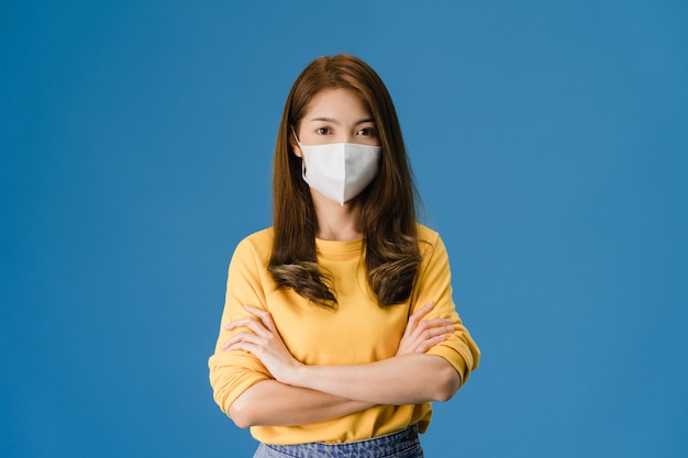 Giovane ragazza asiatica che indossa la maschera medica con le braccia incrociate, vestita in un panno casual e guardando la telecamera isolata su sfondo blu. autoisolamento, allontanamento sociale, quarantena per il virus corona.