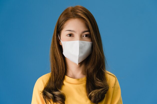 Молодая азиатская девушка в медицинской маске с одетой в повседневную одежду и глядя на камеру, изолированную на синем фоне. Самоизоляция, социальное дистанцирование, карантин для предотвращения вируса короны.