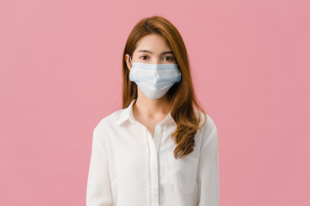 カジュアルな服を着て、ピンクの背景で隔離のカメラを見て医療フェイスマスクを身に着けている若いアジアの女の子。