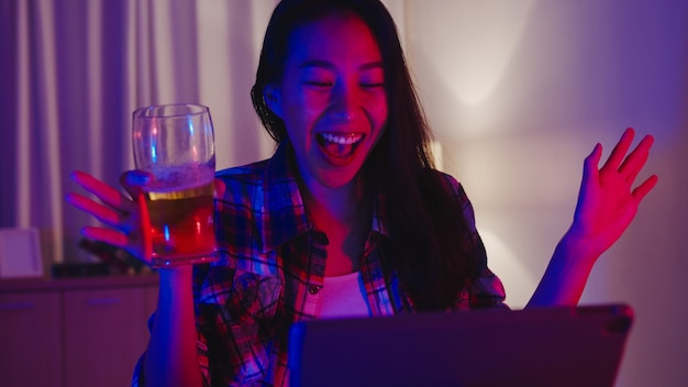 Free photo young asia lady drinking beer having fun happy moment disco neon night party event online celebration via video call in living room at home.