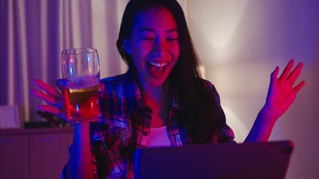 Young Asia lady drinking beer having fun happy moment disco neon night party event online celebration via video call in living room at home.