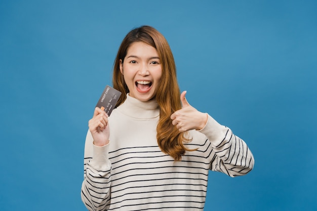 Молодая азиатская леди показывает кредитную банковскую карту с позитивным выражением лица, широко улыбается, одетая в повседневную одежду, чувствуя счастье и стоит изолированно на синей стене