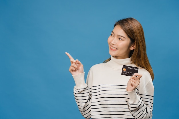 Foto gratuita la giovane donna asiatica mostra la carta di credito con espressione positiva, sorride ampiamente, vestita con abiti casual sentendo felicità e stando isolata sulla parete blu