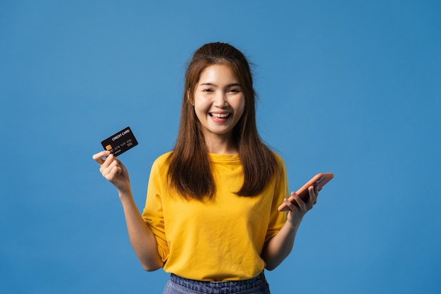 Молодая азиатская леди с помощью мобильного телефона и кредитной карты с позитивным выражением лица, одетая в повседневную одежду и глядя на камеру, изолированную на синем фоне. Счастливая очаровательная рада женщина радуется успеху.