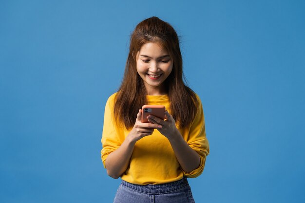 Молодая азиатская леди пользуется телефоном с позитивным выражением лица, широко улыбается, одетая в повседневную одежду, чувствуя счастье и стоя изолированной на синем фоне. Счастливая очаровательная рада женщина радуется успеху.