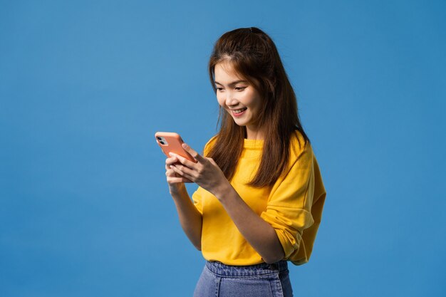 Молодая азиатская леди пользуется телефоном с позитивным выражением лица, широко улыбается, одетая в повседневную одежду, чувствуя счастье и стоя изолированной на синем фоне. Счастливая очаровательная рада женщина радуется успеху.