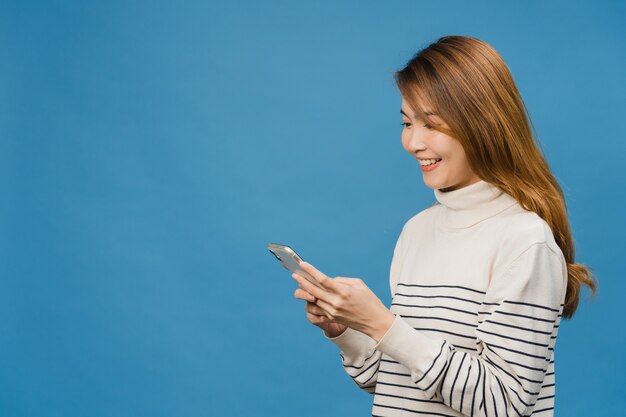 Young Asia lady using phone with positive expression, smiles broadly, dressed in casual clothing feeling happiness and standing isolated on blue wall