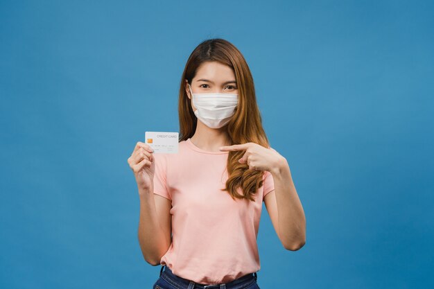 의료용 얼굴 마스크를 쓴 젊은 아시아 여성은 긍정적인 표정으로 신용 은행 카드를 보여주고, 활짝 웃고, 행복을 느끼는 캐주얼 옷을 입고, 파란 벽에 고립되어 있습니다.