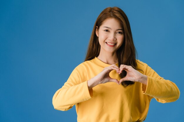 Young Asia lady with positive expression, shows hands gesture in heart shape, dressed in casual clothing and looking at camera isolated on blue background. Happy adorable glad woman rejoices success.