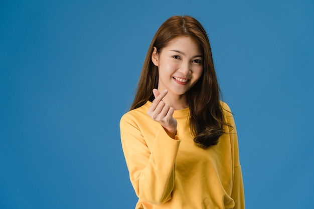 Young Asia lady with positive expression, shows hands gesture in heart shape, dressed in casual clothing and looking at camera isolated on blue background. Happy adorable glad woman rejoices success.