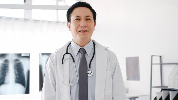 Young Asia male doctor in white medical uniform with stethoscope looking at camera, smile and arms crossed while video conference call with patient in health hospital.