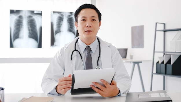 Young Asia male doctor in white medical uniform with stethoscope using computer laptop talk video conference call with patient, looking at camera in health hospital.