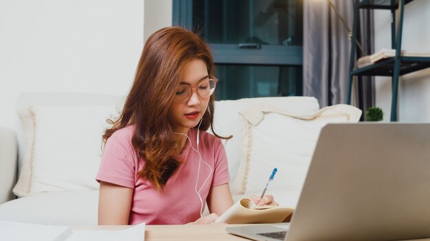Молодая азиатская студентка-подросток носит наушники на уроке дистанционного обучения с онлайн-учителем на ноутбуке в гостиной из дома ночью. Социальное дистанцирование, карантин для предотвращения вируса короны.