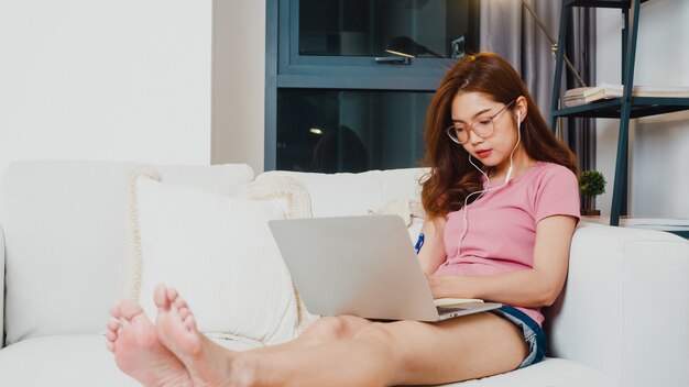 Young Asia teen girl student wears headphones distance learning lesson with online teacher on laptop in living room from home at night. Social distancing, quarantine for corona virus prevention.