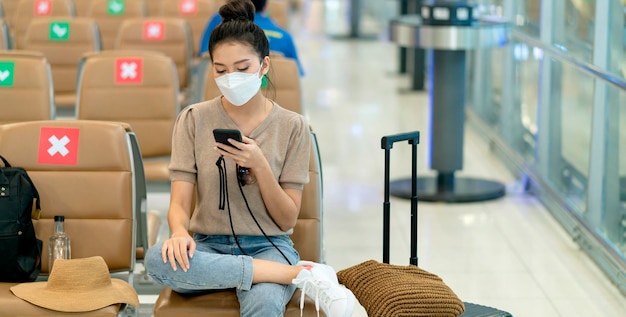 Young asian adult traveller wearing face mask hand hold smartphone sitting with social distancing wait for flight schedule at lobby area at airport terminal safety travel ideas concept