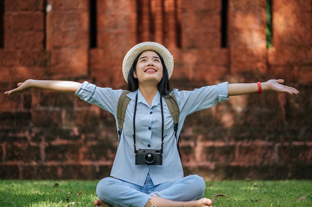 Free photo young asian backpacker female wearing hat smiling while has trip in historic site, she sitting on grass for relaxing and use camera take a photo with happy