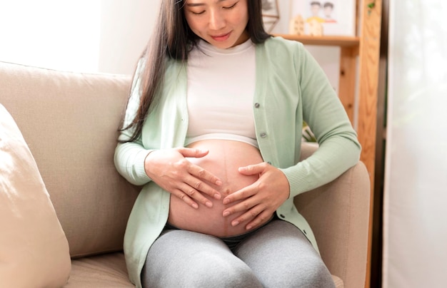 Руки молодой азиатской красивой беременной женщины ласкают живот, сидя дома на диване