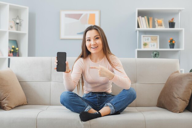 Young asian beautiful woman in home clothes sitting on a couch at home interior showing smartphone pointing with index finger at it looking confident smiling spending time at home