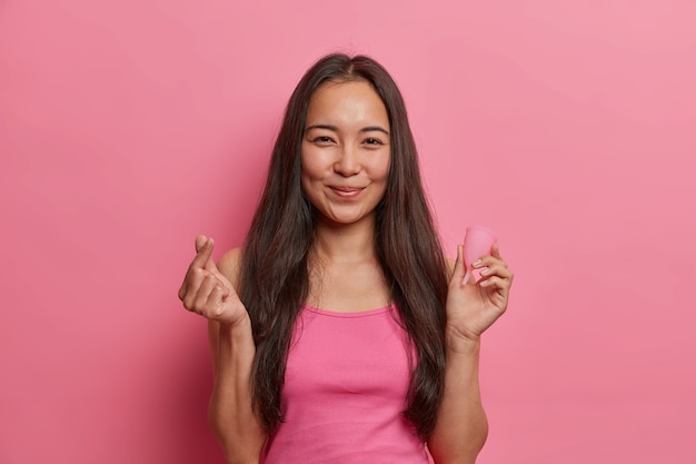 Free photo young asian brunette woman poses with menstrual cup, shows how to use, gives recommendations, makes korean like sign, isolated on pink wall. women health, zero waste alternative and gynecology