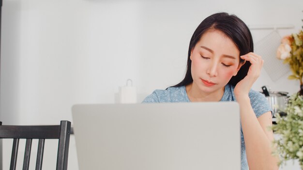 Молодые азиатские записи бизнес-леди дохода и расходов дома. Леди переживает, серьезно, стресс при использовании ноутбука рекордный бюджет, налог, финансовый документ, работающий в современной кухне дома.