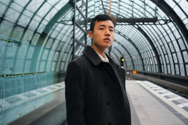 현대 지하철 역에서 카메라를 기다리는 기차를 열심히 보고 있는 코트를 입은 젊은 아시아 사업가