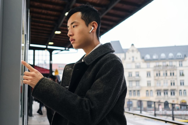 Giovane uomo d'affari asiatico in auricolari wireless che guarda pensieroso il percorso dei trasporti pubblici sulla strada della città