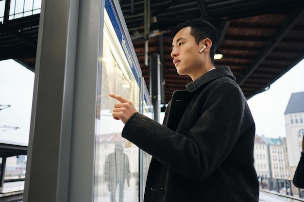 Молодой азиатский бизнесмен в беспроводных наушниках задумчиво смотрит расписание общественного транспорта на станции метро под открытым небом