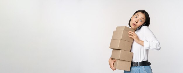 Young asian businesswoman answer phone call talking on mobile while carrying pile of boxes with orders standing over white background