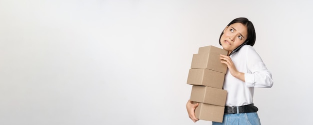 Young asian businesswoman answer phone call talking on mobile while carrying pile of boxes with orders standing over white background