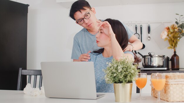 Young Asian businesswoman serious, stress, tired and sick while working on laptop at home. Husband give her glass of water while working hard in modern kitchen at house in the morning .