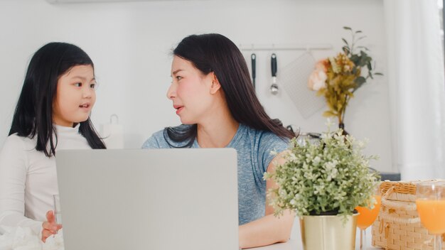 Young Asian businesswoman serious, stress, tired and sick while working on laptop at home. Young daughter consoling her mother who working hard in modern kitchen at house in the morning .