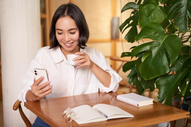 カフェに座ってノートコーヒーを飲み、スマートフォンのビデオチャットを見ているアジアの若いビジネスウーマン