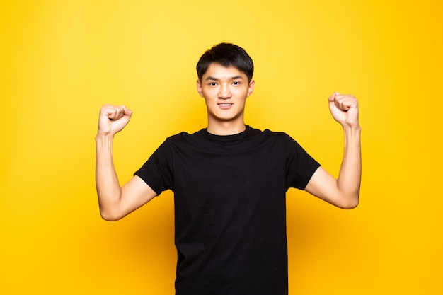 腕を上げると孤立した黄色の壁の上に立って目を開けて驚いて、成功を祝う若いアジアの中国人男性。勝者の概念。