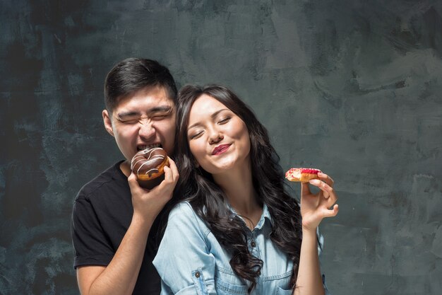 Young asian couple enjoy eating of sweet colorful donut on gray studio background