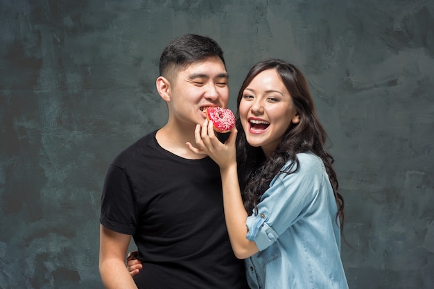若いアジアのカップルは灰色のスタジオで甘いカラフルなドーナツを食べることを楽しむ