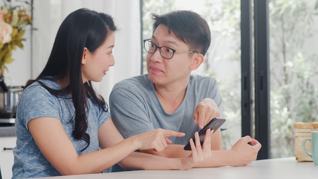 Young asian couple enjoy shopping online on mobile phone at home. lifestyle young husband and wife happy buy ecommerce after have breakfast in modern kitchen at house in the morning .