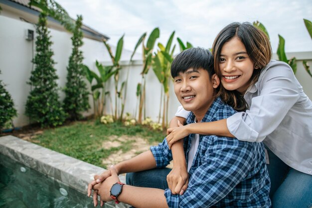若いアジアのカップルは、新しい家のプールの近くで休んでいる間一緒に楽しむ若いハンサムな男は妻の幸せな家族の概念の肩を抱きしめます