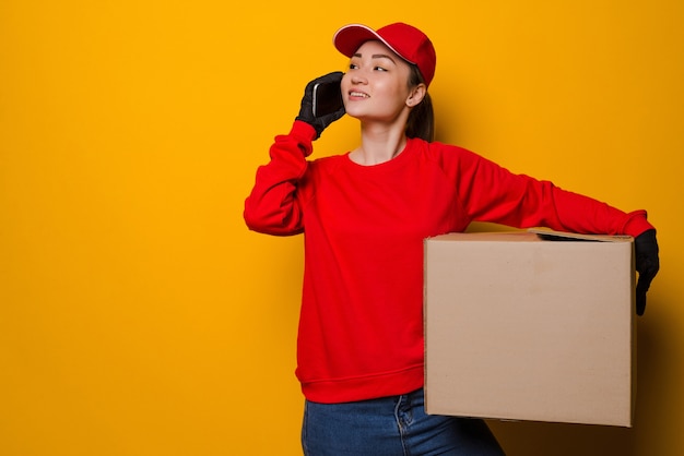 Young asian delivery woman holding box talking on the phone isolated on yellow wall