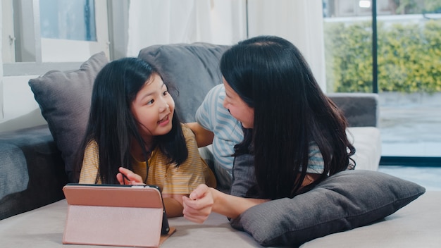 Молодая азиатская семья и дочь счастливые используя таблетку дома. Японская мать отдохнуть с маленькой девочкой, смотреть фильм, лежа на диване в гостиной в доме. Веселая мама и милый ребенок веселятся.