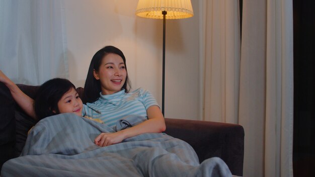 Young Asian family and daughter watching TV at home in night. Korean mother with little girl happy using family time relax lying on sofa in living room. Funny mom and lovely child are having fun.