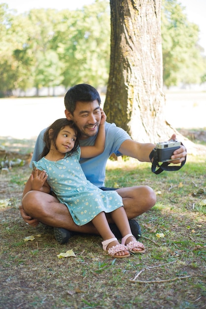 Young Asian father and child taking their photos on camera. Smiling man sitting on ground with little daughter on his knees and taking pictures both looking at camera. Leisure and fatherhood concept