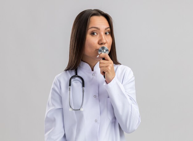 Молодая азиатская женщина-врач в медицинском халате и стетоскопе трогает губы стетоскопом, изолированным на белой стене с копией пространства