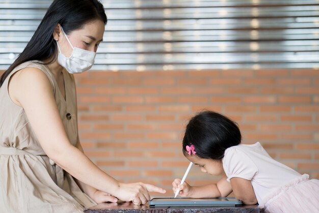 Young Asian female in a facial mask showing her daughter how to write on a tablet