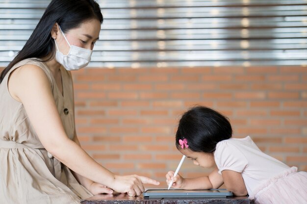 그녀의 딸이 태블릿에 쓰는 방법을 보여주는 안면 마스크를 쓴 젊은 아시아 여성