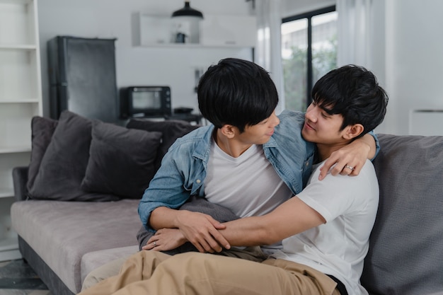 젊은 아시아 게이 커플 포옹과 집에서 키스. 매력적인 아시아 LGBTQ 자부심 남자는 휴식을 취하면서 거실에서 소파에 누워있는 동안 낭만적 인 시간을 함께 보냅니다.