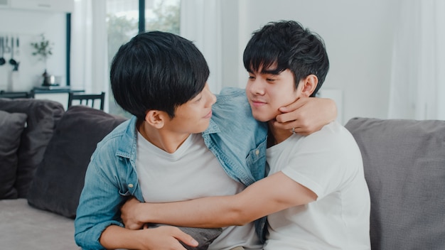 若いアジア同性愛者のカップルが抱擁し、自宅でキスします。魅力的なアジアのLGBTQの誇り男性はリラックスして、リビングルームでソファに横たわっている間一緒にロマンチックな時間を過ごします。
