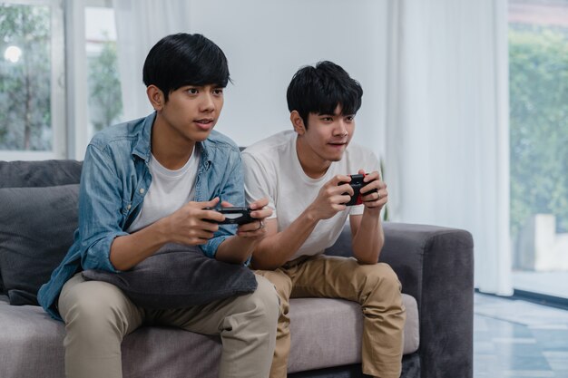 Young Asian gay couple play games at home, Teen korean LGBTQ men using joystick having funny happy moment together on sofa in living room at house.