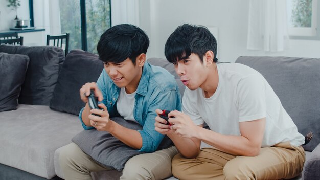 Young Asian gay couple play games at home, Teen korean LGBTQ+ men using joystick having funny happy moment together on sofa in living room at house.