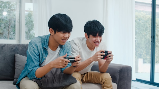 Young Asian gay couple play games at home, Teen korean LGBTQ+ men using joystick having funny happy moment together on sofa in living room at house.