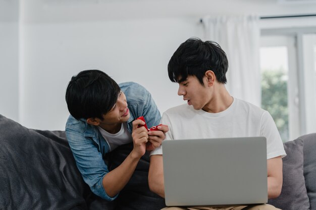 Young Asian gay couple propose at modern home, Teen korean LGBTQ men happy smiling have romantic time while proposing and marriage surprise wear wedding ring in living room at house .